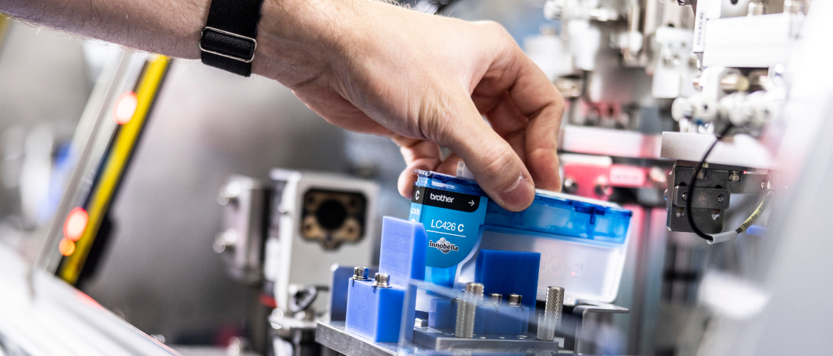 Hand with watchstrap holding the top of a cyan ink cartridge placing it into a machine