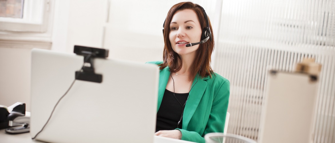 A woman wearing a microphone headset and a green business suit conducts a remote online video conferencing meeting in a home or small office environment. 