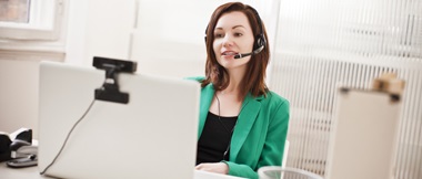 A woman wearing a microphone headset and a green business suit conducts a remote online video conferencing meeting in a home or small office environment. 