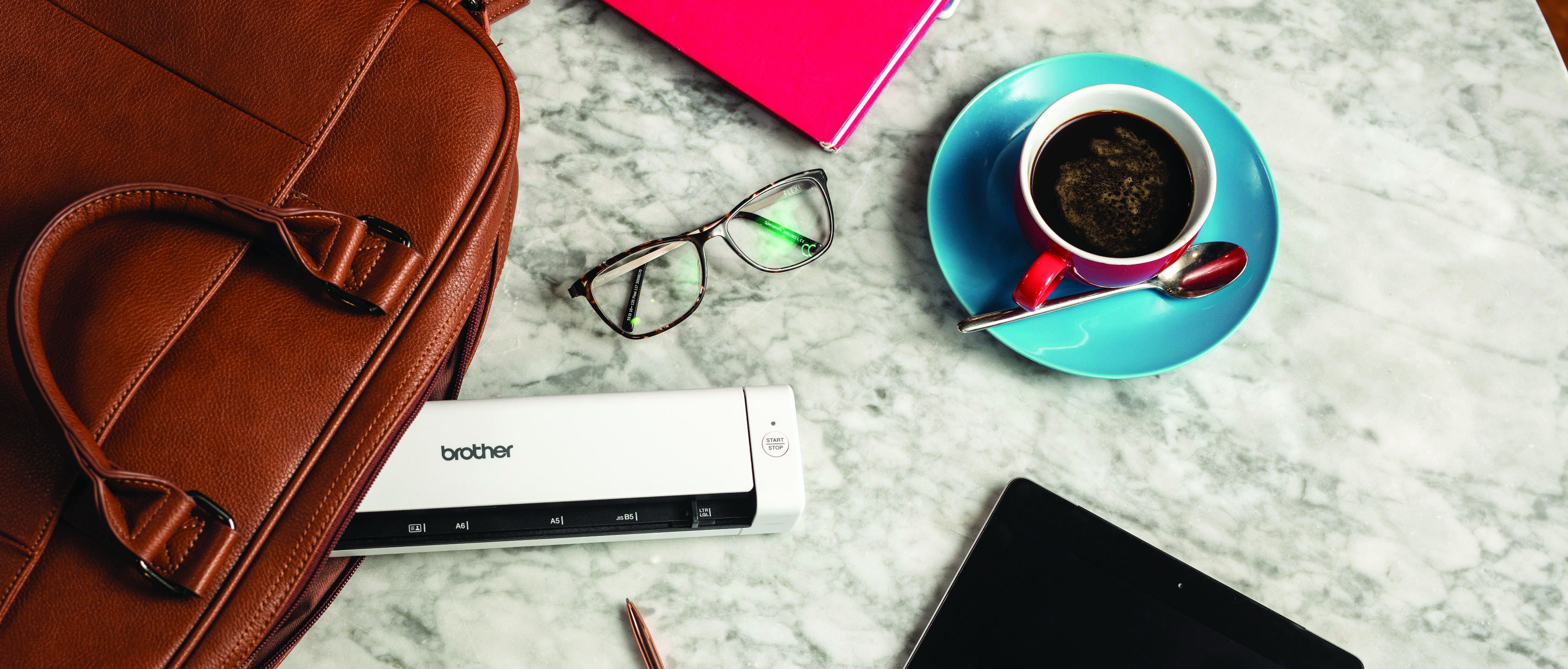 A flatlay of a white Brother portable mobile scanner, turquoise coffee cup and saucer, black ipad, bright pink notebook, brown leather laptop bag, a pair of glasses, a pencil on top of a marble effect table background